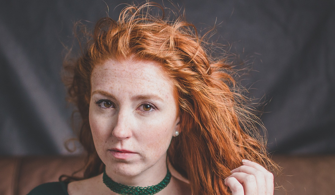 Moira from Meadowsweet. Woman with pale skin, wavey red hair, wearing a green laced top and matching choker.